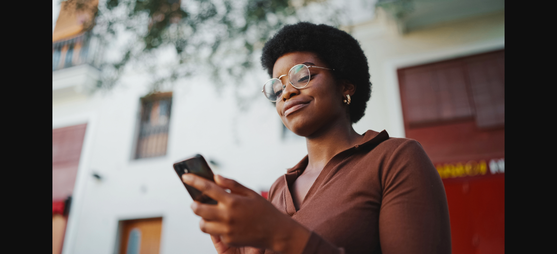 Mulher de óculos sorrindo enquanto usa o celular ao ar livre